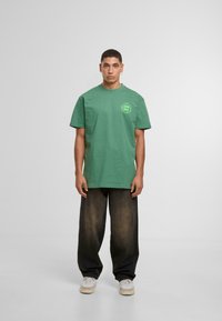Camiseta de algodón verde con mangas cortas, con un escote redondo y un detalle gráfico en el lado izquierdo del pecho. Lleva puesta con pantalones de mezclilla negros holgados.