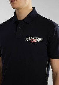 Tummansininen polo-paita, joka on valmistettu hengittävästä kankaasta. Paidassa on klassinen kaulus, kolmen napin lista ja logograafi, jossa on Norjan lipun yksityiskohta.