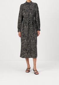 Czarna sukienka w biały wzór paisley, z kołnierzykiem, długimi rękawami, paskiem do wiązania i midi długości. Płaskie czarne sandały.