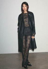 Crni kožni dugi kaput preko prozirne crne čipkaste bluze i hlača s cvjetnim vezom, upotpunjeno crnim cipelama na visoku petu.