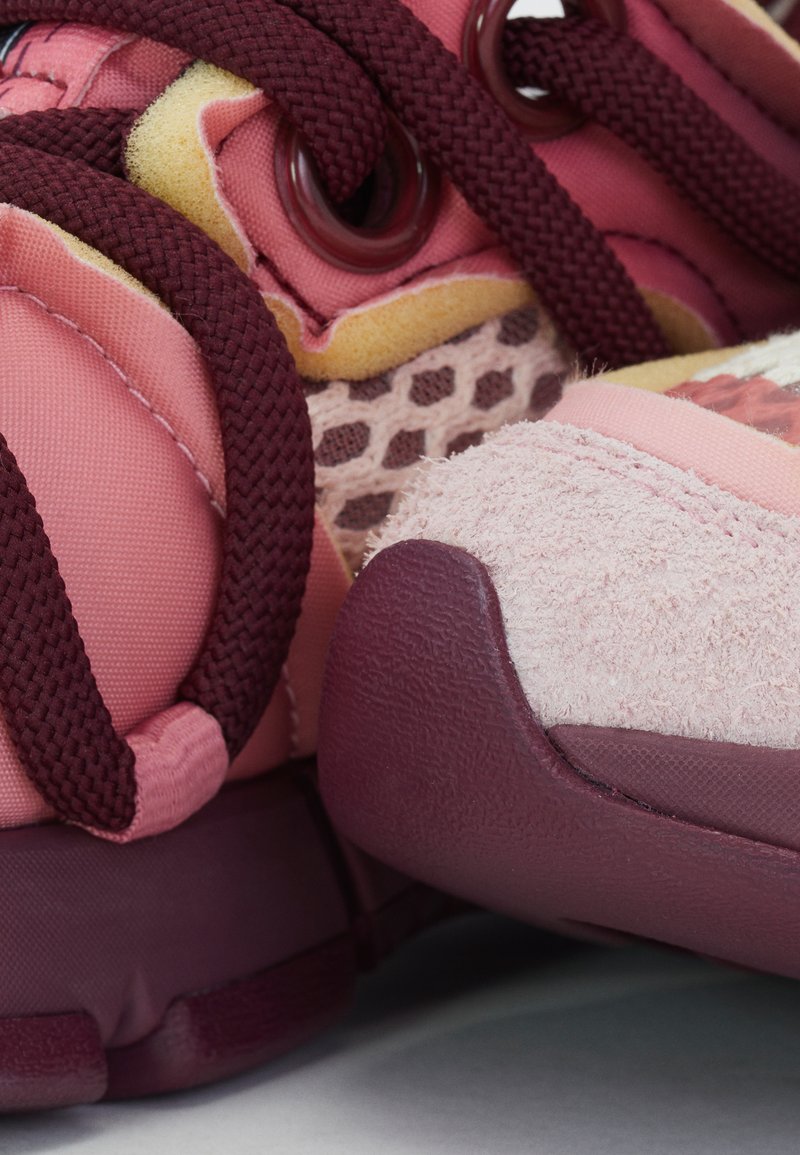 Scarpa da ginnastica rosa con lacci color bordeaux, rete e dettagli in suede; presenta superfici testurizzate e una suola in gomma spessa.