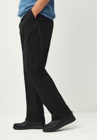 Pantalon noir à coupe droite, avec poches latérales. Associé à des chaussures noires à enfiler avec une semelle épaisse. Pull bleu visible au-dessus.