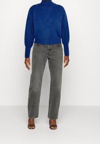 Person, die einen blauen gerippten Rollkragenpullover, ausgewaschene graue Gerade-Jeans und schwarze Sandalen mit offenen Zehen trägt, steht vor einer weißen Wand.