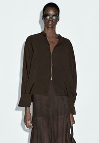 Bruine knoopshirt met een losse pasvorm, voorzien van een kraag en op maat gemaakte naden, gecombineerd met een bruine rok. Model draagt donkere zonnebril.
