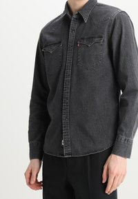 Camicia in denim nero con colletto, maniche lunghe, due tasche sul petto e chiusura frontale con bottoni. Presenta cuciture a contrasto e orlo curvo.