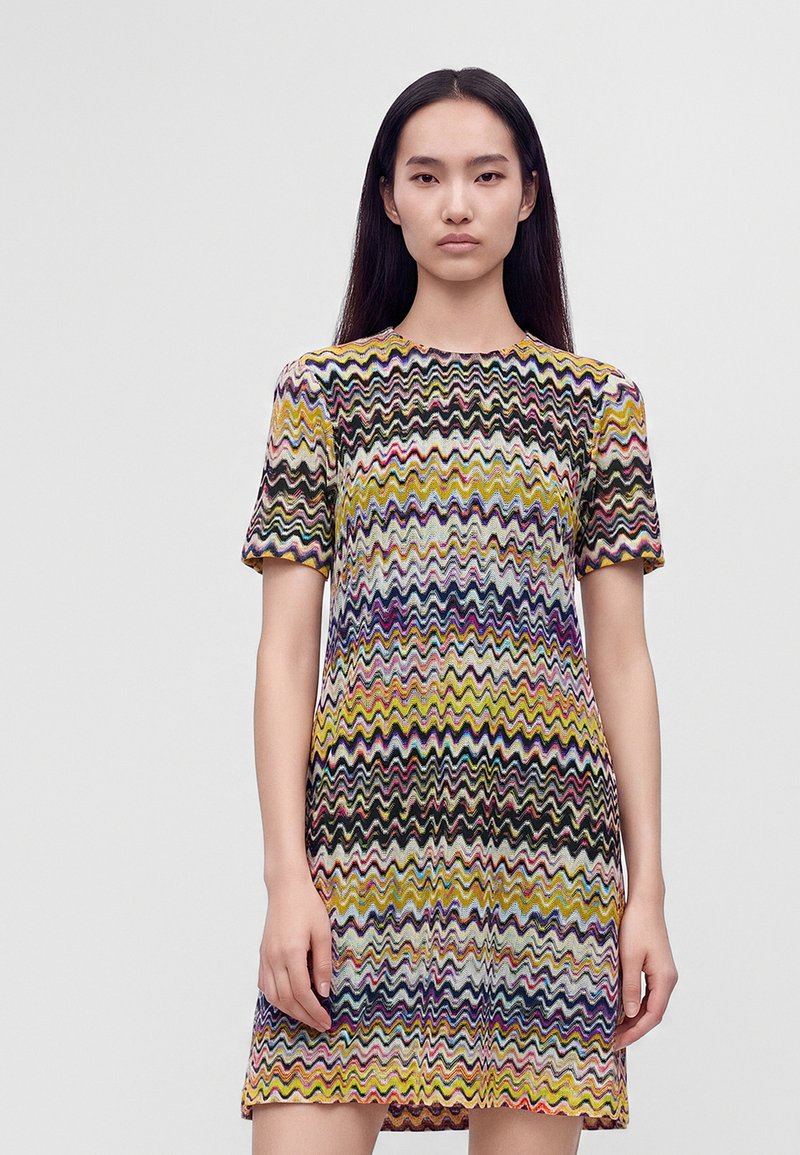 Femme aux longs cheveux noirs portant une robe courte à manches courtes et longueur genou avec un motif zigzag coloré, debout devant un fond uni.