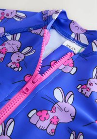 Laste ujumistrikoo erksinisel taustal, millel on korduv lilla jäneste muster roosa aktsentidega. Suumitud pildi peal on nähtav roosa tõmblukk.