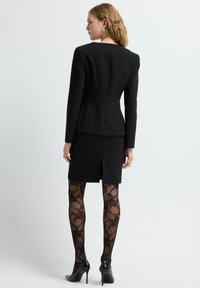 Chaqueta de traje negra ajustada con hombros estructurados, acompañada de una falda ajustada a juego. Medias negras translúcidas con estampado y zapatos de tacón alto.