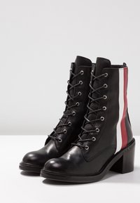 Bottines en cuir noir avec lacets, talons blocs bas, et rayures verticales rouges et blanches sur les côtés, présentées sur une surface blanche.