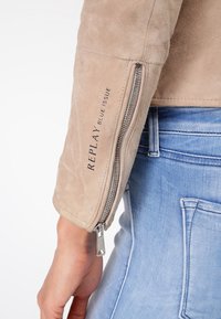 Beige suède jas met een ritsmanchet en zwart "Replay Blue Issue" logo-detail. De jas heeft een gladde, matte textuur.