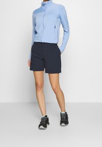 Osoba v světle modré zipové bundě, černých šortkách a černých teniskách stojící s jednou rukou v kapse na jednobarevném pozadí.