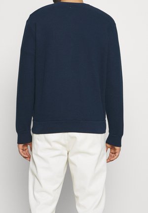 Persona in piedi di spalle, che indossa una maglietta a maniche lunghe blu navy e pantaloni bianchi, su uno sfondo uniforme.