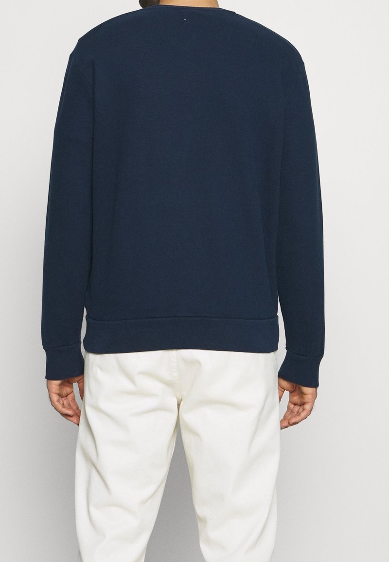 Person som står vänd bort med ryggen mot kameran, iförd en marinblå långärmad skjorta och vita byxor framför en neutral bakgrund.