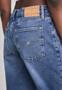 Blå denimjeans med hög midja, med en tan läderetikett märkt "TOMMY JEANS" och bakfickor med subtila sömdetaljer.