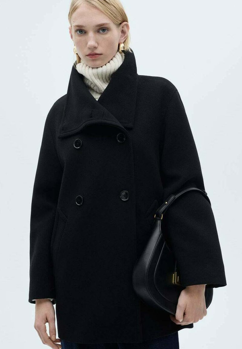 Black double-breasted wool coat with wide collar, large buttons, and side pockets, paired with a white ribbed turtleneck sweater and black purse.