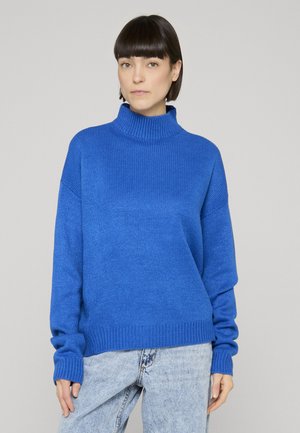 Vrouw met kort donker haar, gekleed in een felblauwe coltrui en lichtgebleekte spijkerbroek, tegen een effen achtergrond.