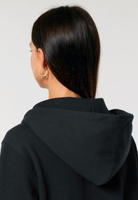 Fekete kapucnis pulcsi széles kapucnival, puha anyagból és látható varrási részletekkel. Arany fülbevaló kiemeli a modell profilját.