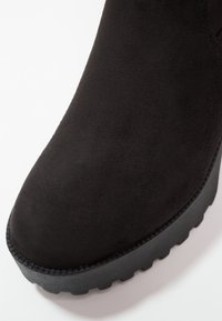 Botín de ante negro con puntera redondeada, superficie texturizada y suela de goma negra gruesa con acentos estriados.