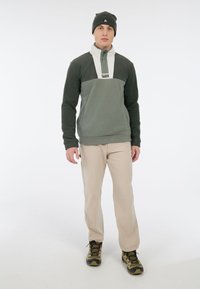 Fleece pullover in donkergroen en roomkleur, met een kwart-ritssluiting. Gecombineerd met beige broeken en groenachtig bruine wandelschoenen, aangevuld met een bijpassende beanie.