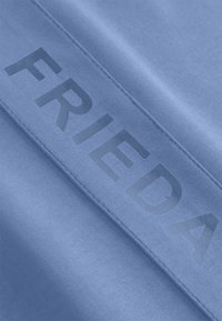Lichtblauwe stof met een gladde textuur, gekenmerkt door een prominente, licht glanzende "FRIEDA" die verticaal aan één zijde is gedrukt.