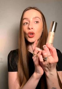 Femme aux longs cheveux bruns tenant et présentant un produit cosmétique MAC avec un bouchon argenté, portant plusieurs bagues aux doigts.