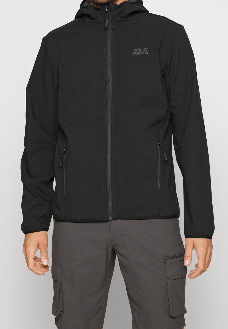 Homme portant une veste zippée noire Jack Wolfskin et un short cargo gris, debout les bras détendus le long du corps.