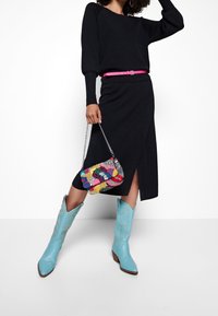 Pull noir en maille à manches longues, jupe portefeuille noire, sac à main vibrant orné de perles, et bottes de cowboy bleu clair avec broderies décoratives.