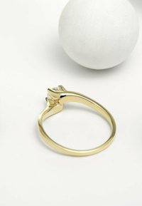 Goldring mit polierter Oberfläche, der ein drehendes Design und einen zentralen klaren Stein aufweist, vor einem glatten weißen Hintergrund.