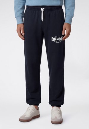 Personne portant un pantalon de survêtement noir Champion avec un cordon blanc et des baskets blanches sur un fond blanc uni.