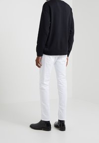 Person i svart långärmad tröja, vita slim-fit jeans och svarta läderankelboots står i ett enkelt vitt utrymme.