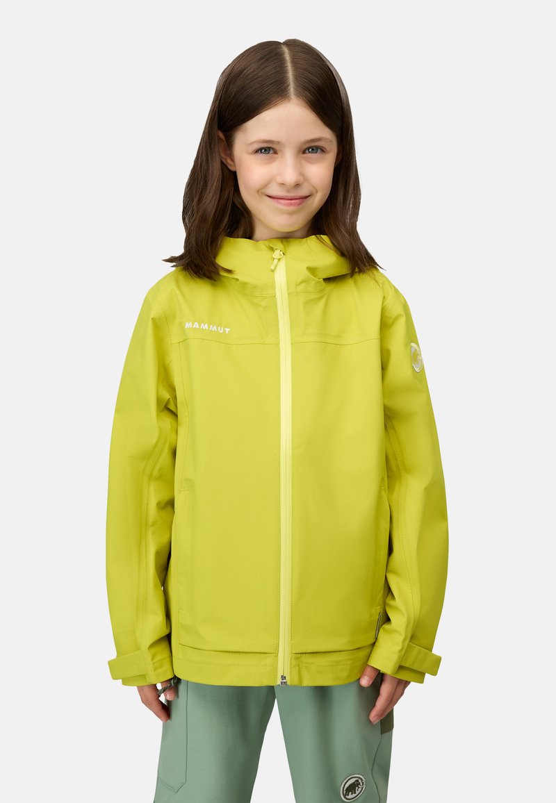 Pige med brunt hår iført limegrøn vandtæt jakke og grønne udendørsbukser, smilende mod en ensfarvet baggrund.