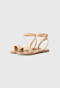 Beige flate sandaler med to brede remmer over tærne og ankelen, med en spenne og kjededetalj, satt på en flat såle.