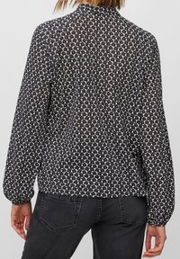 Blusa nera con un piccolo motivo floreale bianco, maniche lunghe e colletto alto. Tessuto trasparente con una vestibilità comoda e polsini arricciati.
