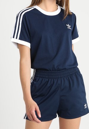 Vrouw draagt donkerblauwe Adidas sportkleding met witte strepen op de mouwen en shorts, staand tegen een effen achtergrond.