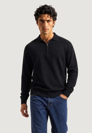 Uomo con capelli ricci e scuri che indossa un maglione nero a maniche lunghe con cerniera e jeans blu, in piedi con una mano in tasca.