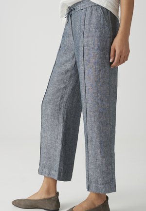 Frau trägt graue Leinenhose mit Kordelzug und Seitentaschen sowie graue flache Schuhe, gezeigt von der Mitte des Oberschenkels bis zu den Füßen vor einfachem Hintergrund.