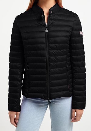 Veste matelassée noire avec fermeture éclair frontale et poches zippées, portée par une personne en jean bleu clair, montrée du cou aux hanches.