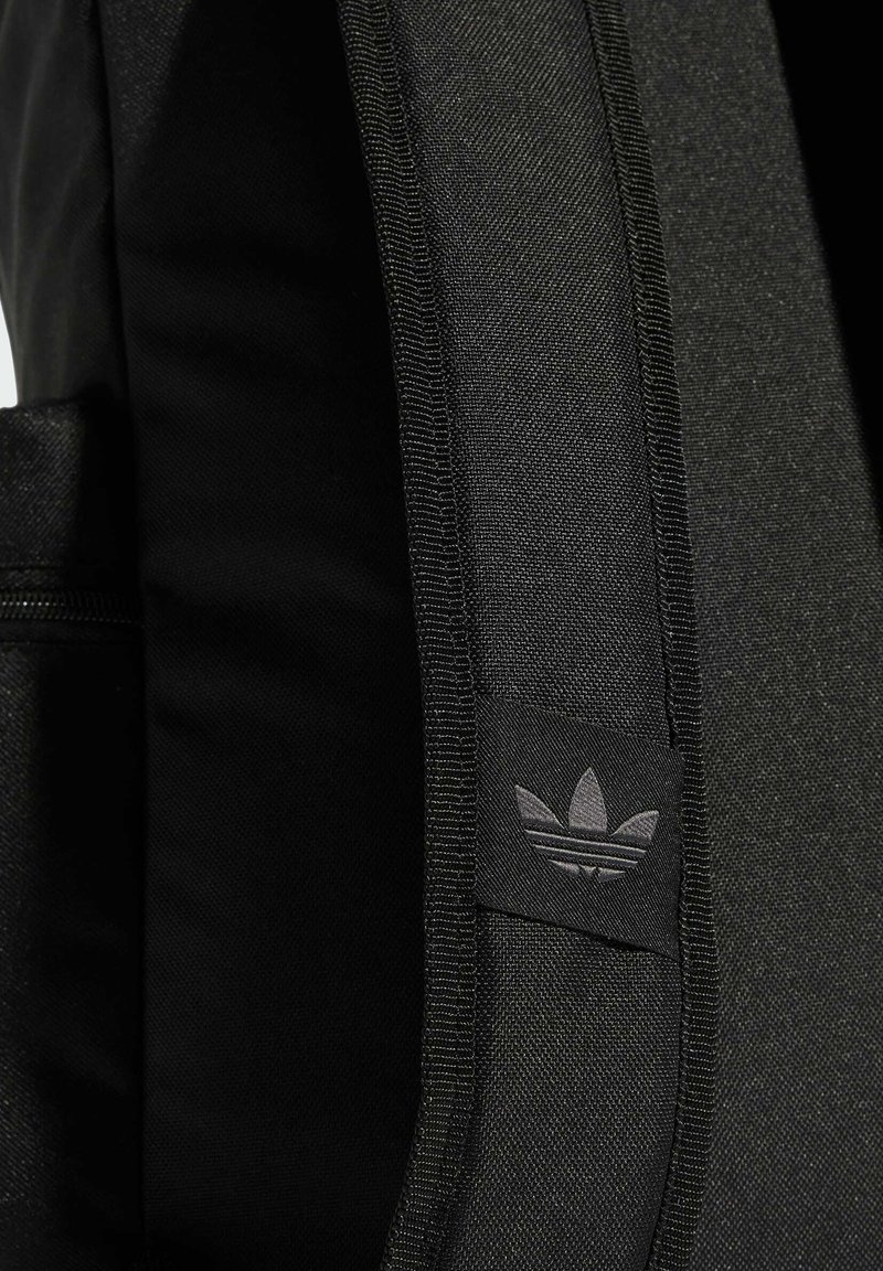 Sac à dos noir avec une texture lisse, arborant une discrète étiquette de logo noire et une poche zippée sur le côté. Détails de couture renforcée.