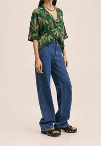 Grünes kurzärmliges Hemd mit goldenen Blättern, kombiniert mit hellblauen, hochtaillierten, geraden Jeans und schwarzen Plateausandalen mit Nieten.