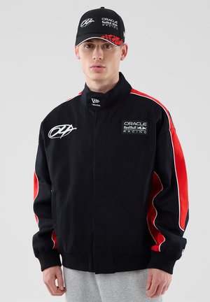 Mladý muž v čierno-červenej bunde Oracle Red Bull Racing a zodpovedajúcej čiapke stojaci na jednofarebnom sivom pozadí.