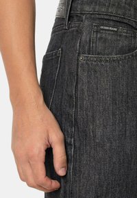Jeans in denim nero con vestibilità slim, dotati di una logo patch visibile sulla cintura e tasche cucite. Il tessuto presenta una sottile texture.