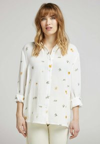 Chemise blanche à boutons avec broderie florale en jaune et vert, avec des manches retroussées et un tissu texturé.