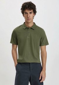 Olijfgroene polo van zacht stof met een kraag, drie knopen en korte mouwen; het model draagt het met een marineblauwe broek.