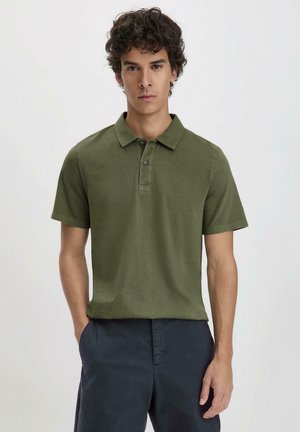 Olivgrünes Polohemd aus weichem Stoff mit Kragen, Dreiknopfleiste und kurzen Ärmeln; das Modell trägt es mit navyblauen Hosen.
