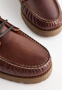 Brune læder loafers med hvid stikning, gummisåler og en rund tå. Har snørrebånd og samlet detalje omkring anklen.