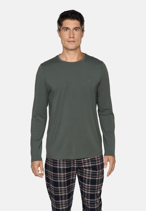 Man wearing a dark green long-sleeve shirt and black, white, and red plaid pants standing against a plain light background.