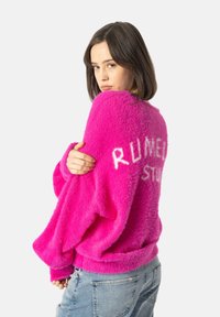Maglione fuzzy rosa brillante con spalle scese, con testo bianco "RUNNER" sul retro e polsini a costine. Indossato sopra jeans blu.