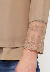 Top beige à manches longues avec poignet en dentelle transparente ornée de broderies florales, porté avec un jean bleu.
