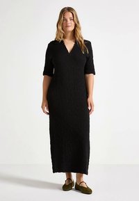 Vestido midi preto com padrão texturizado, decote em V e mangas até ao cotovelo. Modelo usa sapatilhas verdes oliva.