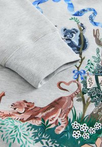 Grijze sweatshirtmouw over een stofafdruk met een uitrekkende tijger, een klimmende koala aan een boom, planten, bloemen en een blauwe strik met de tekst "Hoop &".
